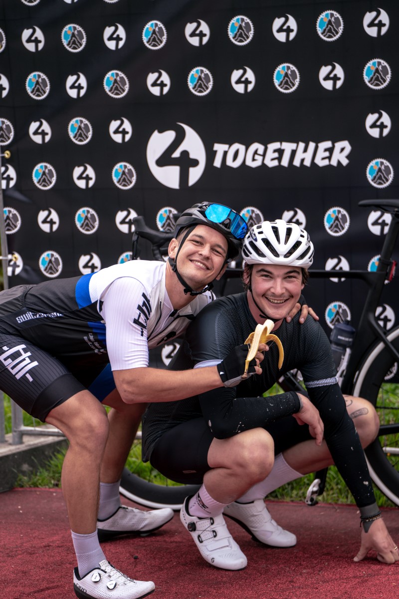 Zwei junge Männer im Fahrradtrikot und mit Helm posieren in wiziger Pose vor dem Plakat mit dem Schriftzug von 24together.